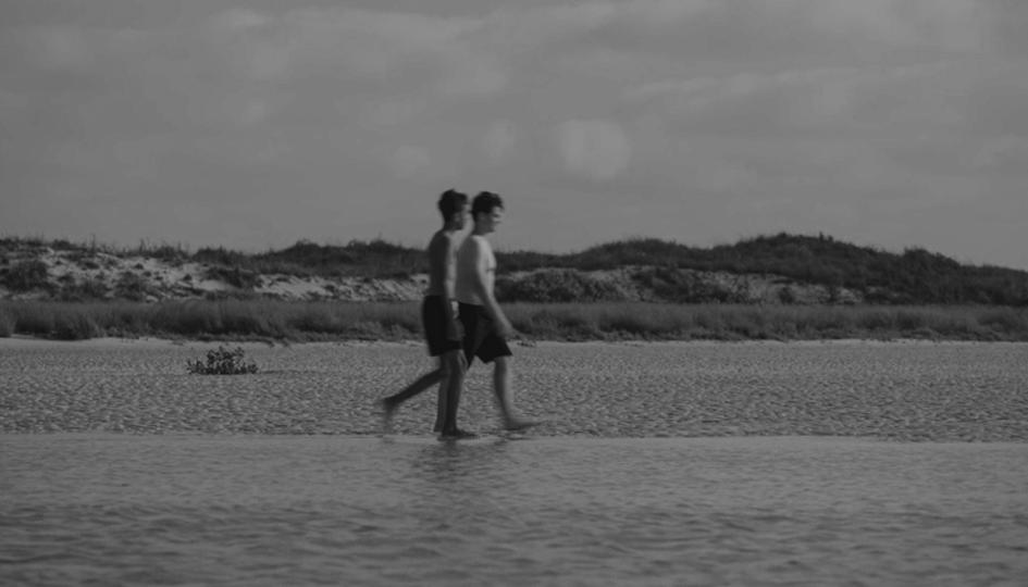 Black and white still of two boys walking across a beach