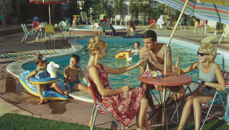 A vintage coloured photo of three people sitting under a pool umbrella at a small round table while their kids play in a motel pool behind them.