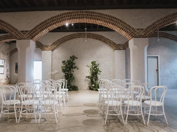 Chairs and flowers are arranged for a wedding in a large room with limestone walls and an arched ceiling 