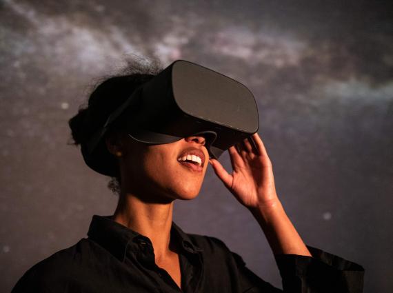 A woman wearing a VR headset stands in front of a starry projected backdrop with an expression of wonder