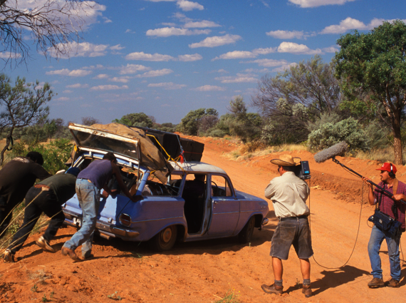 3 people push an old, blue car bogged on the side of a bush track, while 2 camera people film.