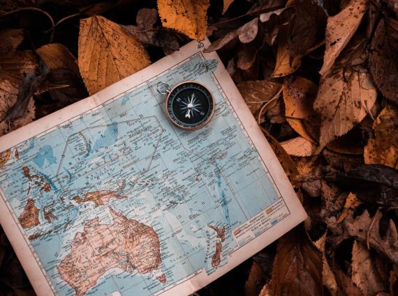 THis image shows an old paper map of Australia laying on a bed of leaves. It also has a compass in the top right hand corner of the map.