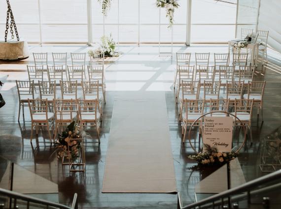 A high-angled photograph looking down a flight of narrow stairs towards two aisles of white chairs in front of a floral arch and large, brightly-lit window.