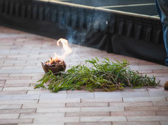 A small pile of green leaves and branches next to a small brown fire receptive with bright yellow flames on a tiled outdoor floor.