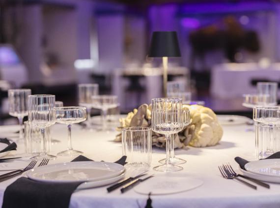 A selection of art deco styled glasses black cutlery and black napkins artfully arranged on a white clothed table in an event space dramatically lit in purple lighting.