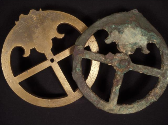 Examples of bronze astrolabes. One was protected by concretion while the other was exposed to changing conditions on the sea bed. One is shiny and metallic, whereas the other looks corroded. Both are the same circular shape, with a plus-shaped cross through the middle.