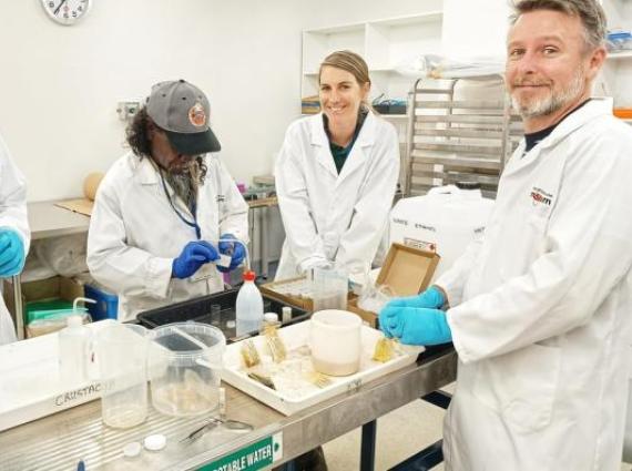 A group of 4 people wearing white coats stand around a bench examining scientific obects