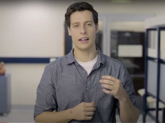 A person with short hair stands in a laboratory space, wearing a light blue long sleeved shirt.