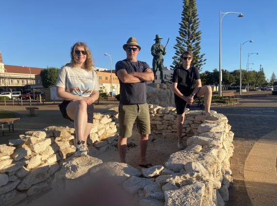 Three people stand on a circular stone structure with a statue and a tree in the background.