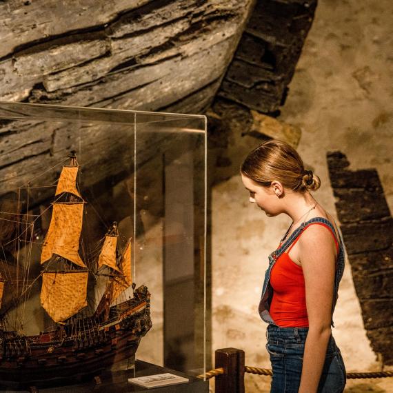 A person stands in a museum gallery, examining a model ship in a showcase