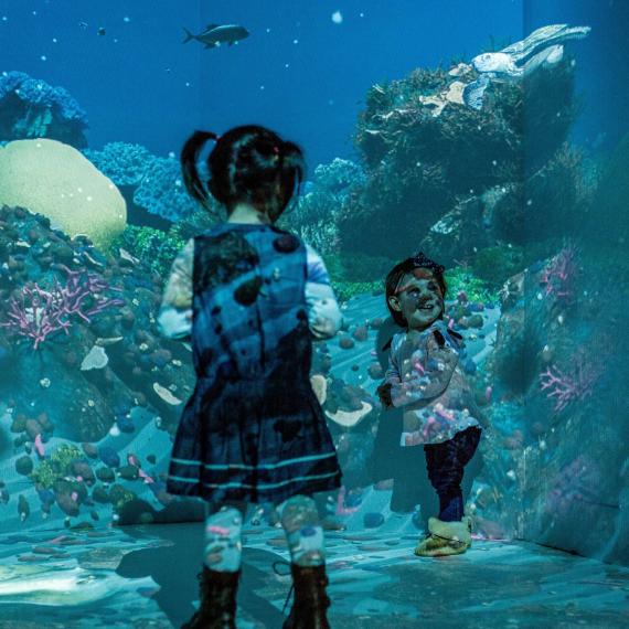 Two young children play in front of a projection wall covered with fish, coral and other underwater life