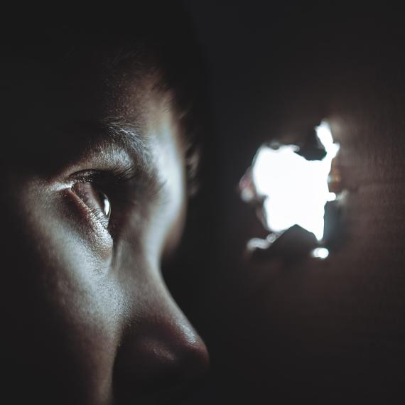 A close up view of a child in the dark peering through a small brightly lit hole in a wooden wall