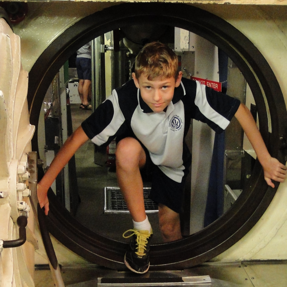 A boy dressed in school uniform climbs through a small hatch door in a submarine