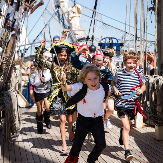 A group of children dressed as pirates rush along the desk of a sailing ship, shouting and waving toy swords