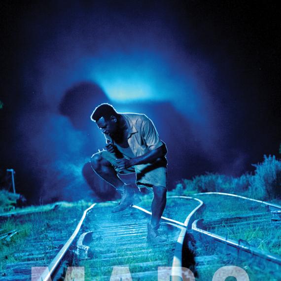 Pictured is a blue tined image of a man in a shirt and shorts with one leg lifted up and head tilted downward with hands clenched close to their chest. They are standing in between two train tracks as the tracks converge. It is dark/night and there is a bright light behind the person 