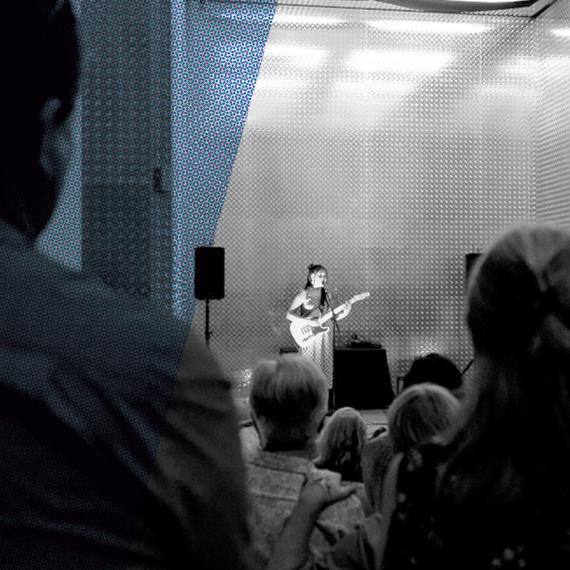 A lone singer stands in the lift entrance with a guitar, a blue strip running along the left side of the black and white image