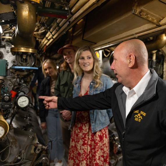 Submarine tour guide gestures inside submarine, three people look on