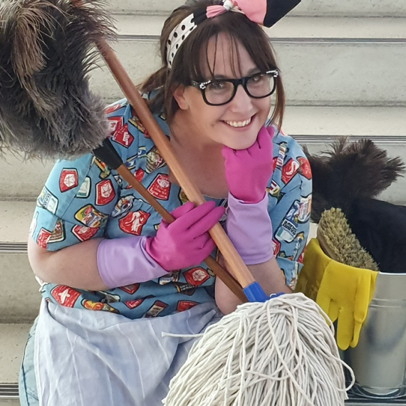 A woman crouches, smiling, while holding cleaning equipment. She wears glasses and pink cleaning gloves, with brown hair pulled back and a ribbon around her head holding her hair in place. 