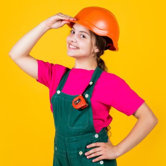 Little girl dressed as a construction worker with tape measure wearnig a orange helmet, pink shirt, green overalls infront of a yellow background. 