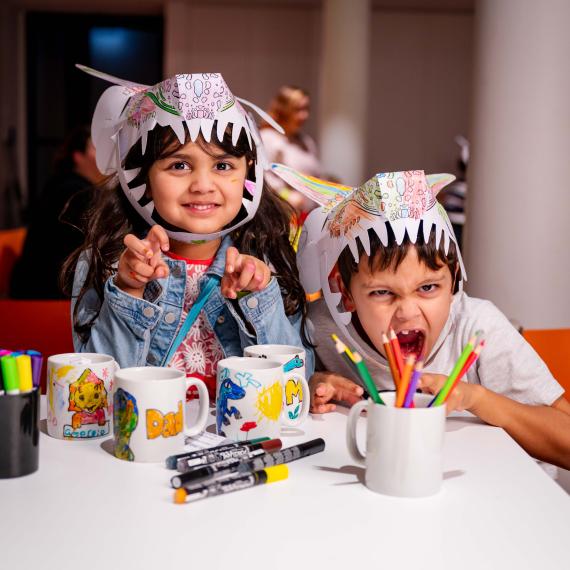Image of two children roaring at the camera wearing paper dinosaur hats