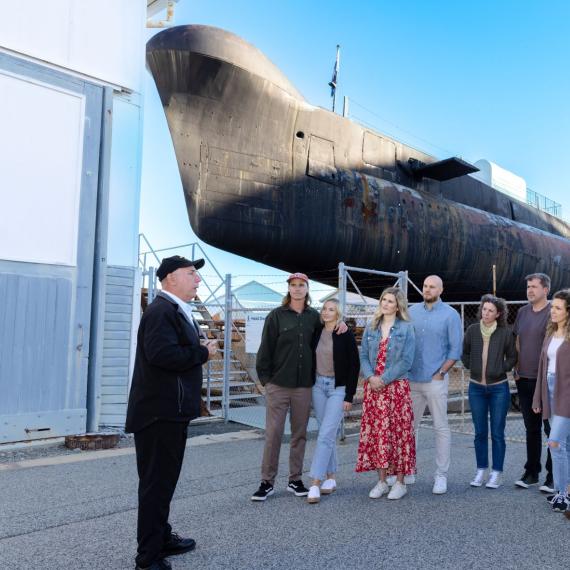 A submarine volunteer speaks to a group of visitors in a semi circle, standing in front of the submarine HMAS ovens
