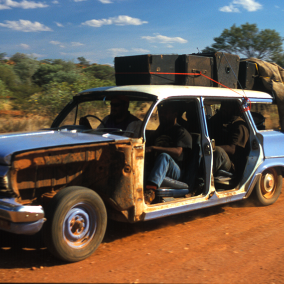 An old blue car with luggage on the top and four passengers drives over red dirt with blue skies overhead in the Australian outback.