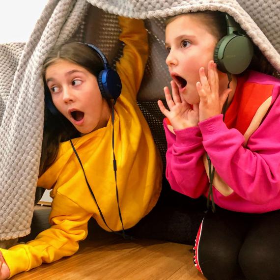 two girls in birght jumpers with headphones on shout and play under a blanket