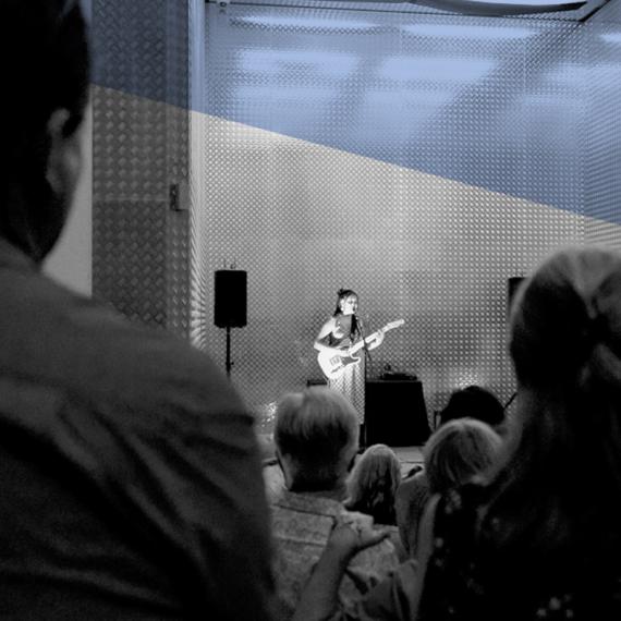 Woman standing in a lift, singing into a microphone holding a guitar