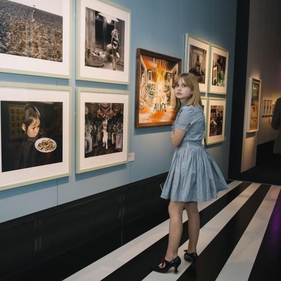 Girl dressed as Alice in Wonderland looks at a blue wall covered in Wonderland drawings and photos. 