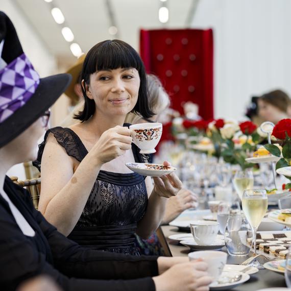 People sitting at a long table holding teacups. The table is set with food and drink items