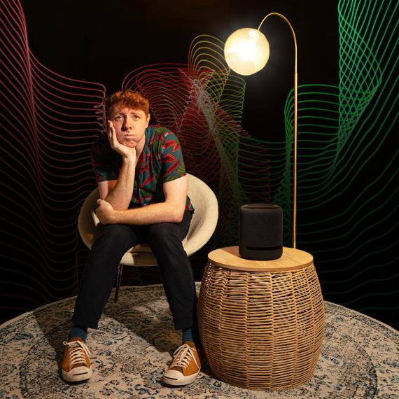 A person with a brown shirt, black jeans and orange sneakers sits on a sofa next to a light infront of LED lights