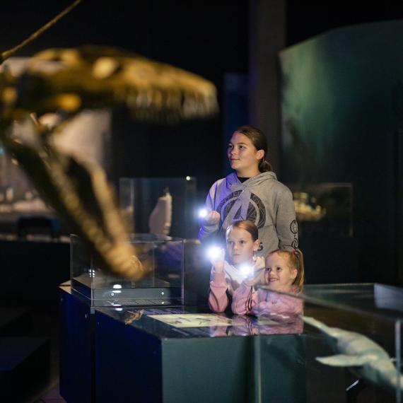 Three young people holding torches in a darkened exhibition with a skeleton in the foreground 