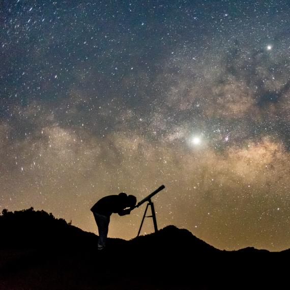 A silhouette of a person gazing through a telescope, in front of a vibrant night sky filled with stars, and deep blue and gold tones