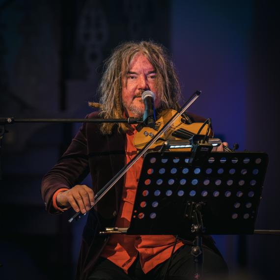 Musician Rupert Guenther sitting on a stage before an audience playing a violin. Behind him is a large screen with a projection of the West Australian landscape.