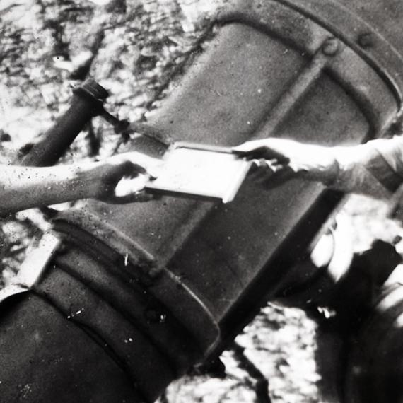 black and white image showing two hands holding a photographic slide. one hand on the right and other on left of photograph edge to indicate they belong to different people and are passing the slide to one another. A large drum of a telescope with viewing device is in the background. 