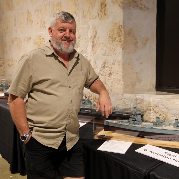 A person with a beard stands smiling in front of model warships display
