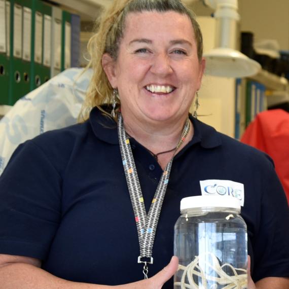 A person with curly hair, wearing a navy-coloured shirt with a logo reading 'CORE" and a patterned lanyard smiles into the camera as they hold a jar containing a white sea star. 