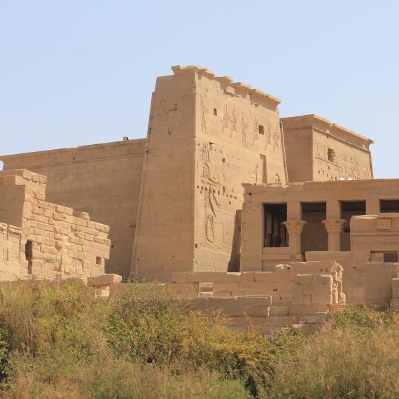 A land sandstone temple positioned in desert-like area with dry green shrubbery stands against a clear blue sky, engraved with large images of people in an Ancient Egyptian style