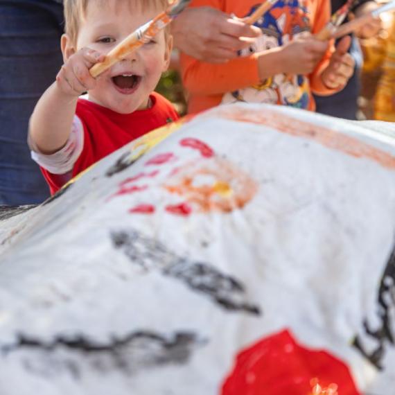 In the photo, a cheerful child is holding a paintbrush with a big smile on their face. They are looking attentively at a sheet adorned with the vibrant colors of the Aboriginal flag.igianl flag