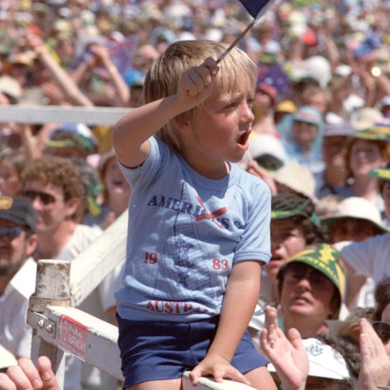 Image of a small boy waving a Western Australian flag in a crowd, celebrating the America's Cup win by Australia II on Perth Esplanade, 1983