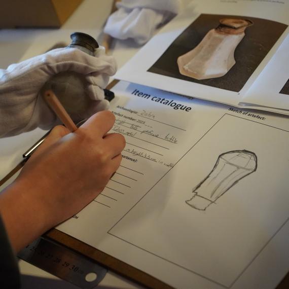 This image shows a close up of someones hand sketching and recording a glass object they uncovered. They are also making some notes on the object to record specific details as an archaeologist would. 