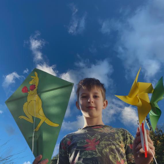 A child of approx 10 years is smiling and holding green kite with a yellow kangaroo and in the other hand they hold yellow and green spinning wheels. A blue sky is in the background