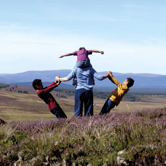 A mother and three children stand and look out onto the moor. One is on her shoulders, the other two hold an arm each and lean away from her.