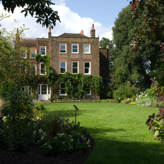 Flat green lawn area surrounded by English garden and tall trees leading towards a three story old brown brick large house
