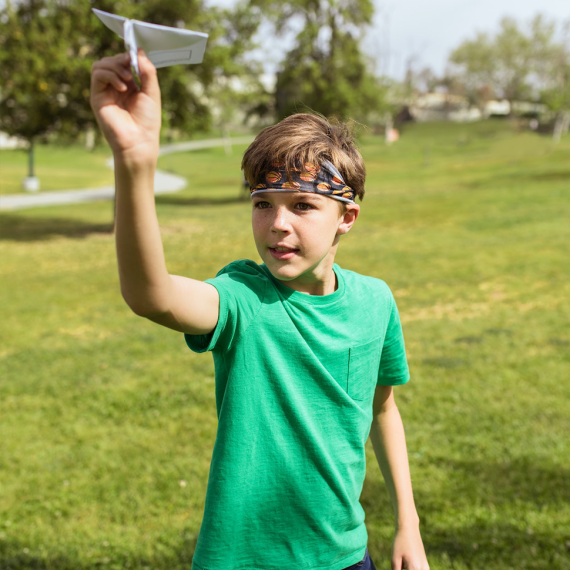 a child stands in a park with a green tshirt on and a headband. they hold a paper plane in one hand and aim to throw it