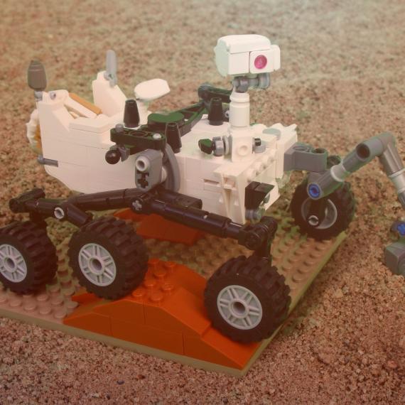 a small planeta lander is made out of lego block pieces mostly white pieces and has six sets of wheels driving over terrain which hs rocky and sandy. there is a slightly green and red tinge to the picture