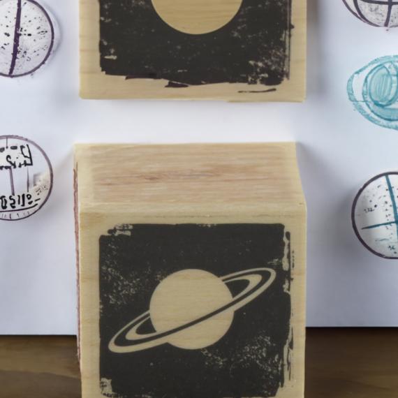 a woodern block stamp of saturn with another planet orb stamp just behind it with a page of a few planet stamps