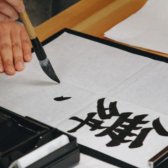 A hand holds a paintbrush which paints with black ink on a white page. the writing is calligraphy of Japanese language characters