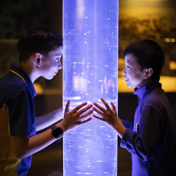 The image shows tow boys standing either side of a large plastic tube filled with a liquid with air bubbles going through it. It looks purple in colour