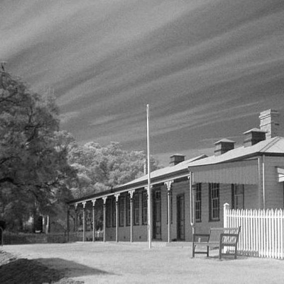 Black and white photos of an old style weather board clad building with a front verandah, and next door is a white picket fence.  On the other side of the building are tall trees.